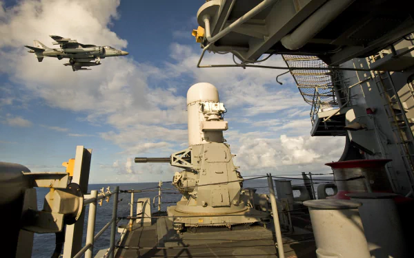 HD desktop wallpaper featuring a McDonnell Douglas AV-8B Harrier II military jet flying over a naval ship deck under a partly cloudy sky.