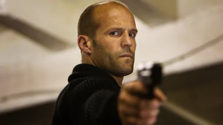 HD PC wallpaper: close-up of a bald action actor aiming a handgun in a tense movie scene, Hummingbird movie–style background.