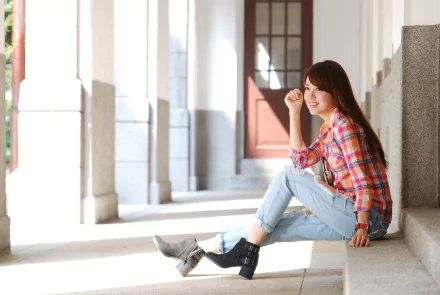 HD desktop wallpaper featuring a Taiwanese woman named Xiǎo Dàn sitting on steps in a bright, open corridor, wearing a plaid shirt and jeans, smiling contentedly.