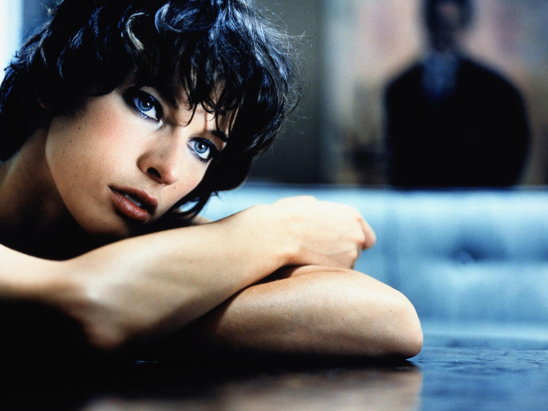 HD PC desktop wallpaper featuring a close-up of celebrity Milla Jovovich with striking blue eyes and short dark hair, resting her arms on a reflective surface.