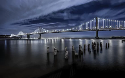 HD PC desktop wallpaper: nighttime view of the man-made San Francisco Bay Bridge spanning calm water, lights reflecting on the bay with weathered pilings in the foreground.
