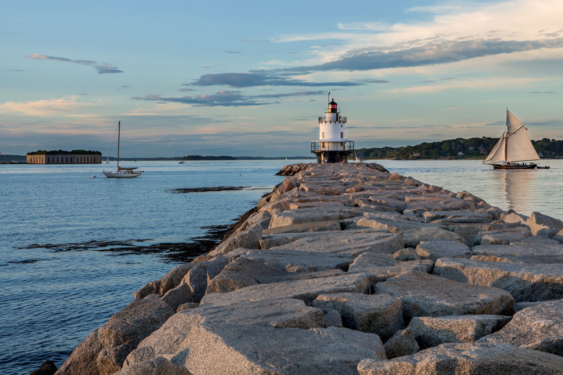 4K Ultra HD PC desktop wallpaper of a man-made lighthouse on a rocky jetty with sailboats on calm coastal waters at sunset.