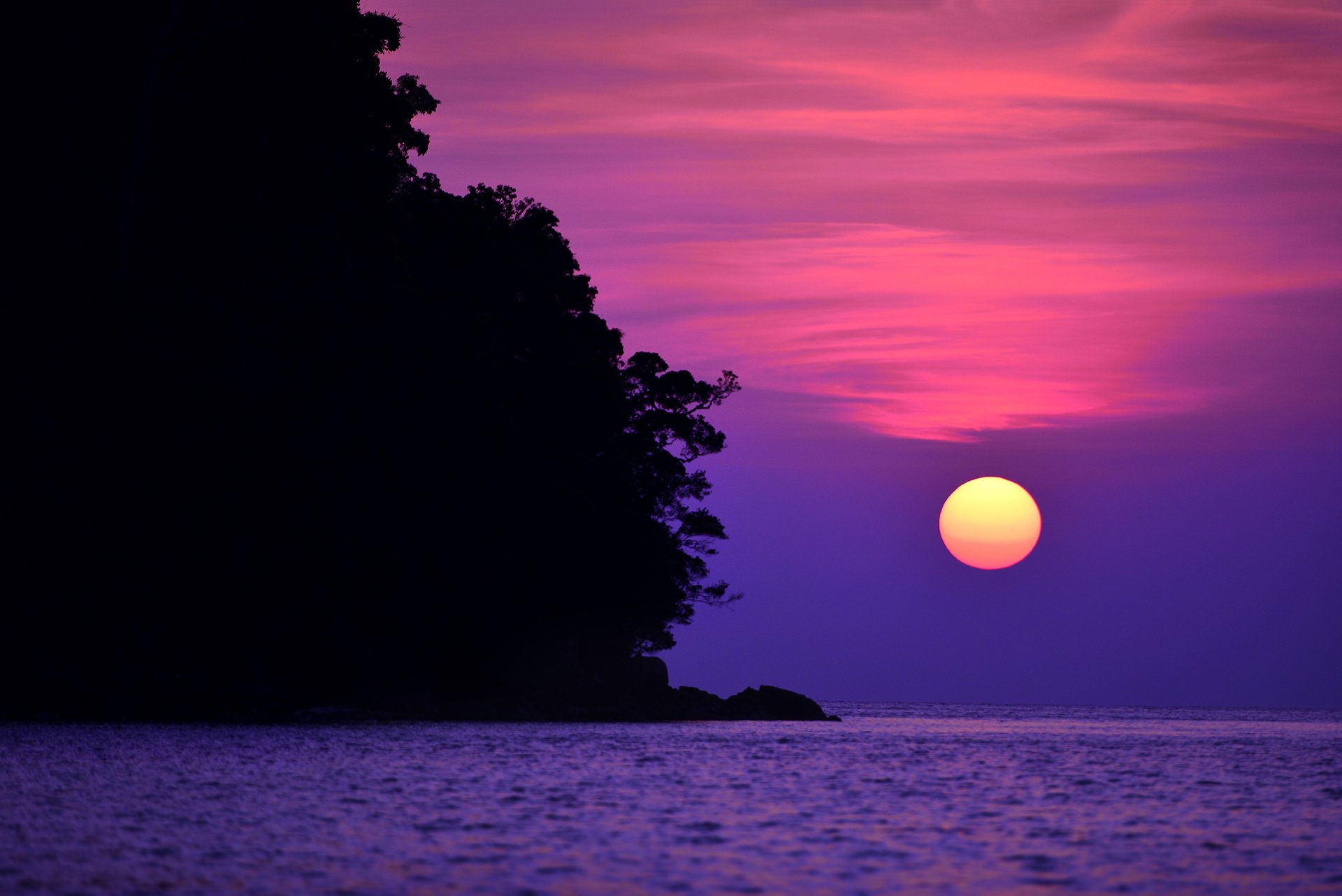 A stunning sunset over the sea in Thailand, with vibrant purples and oranges reflecting on the water, framed by the silhouette of trees. A serene nature scene for an HD desktop background.
