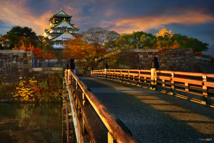 A scenic view of Osaka Castle surrounded by vibrant fall foliage, with a wooden bridge leading to the majestic structure against a backdrop of a colorful sky.