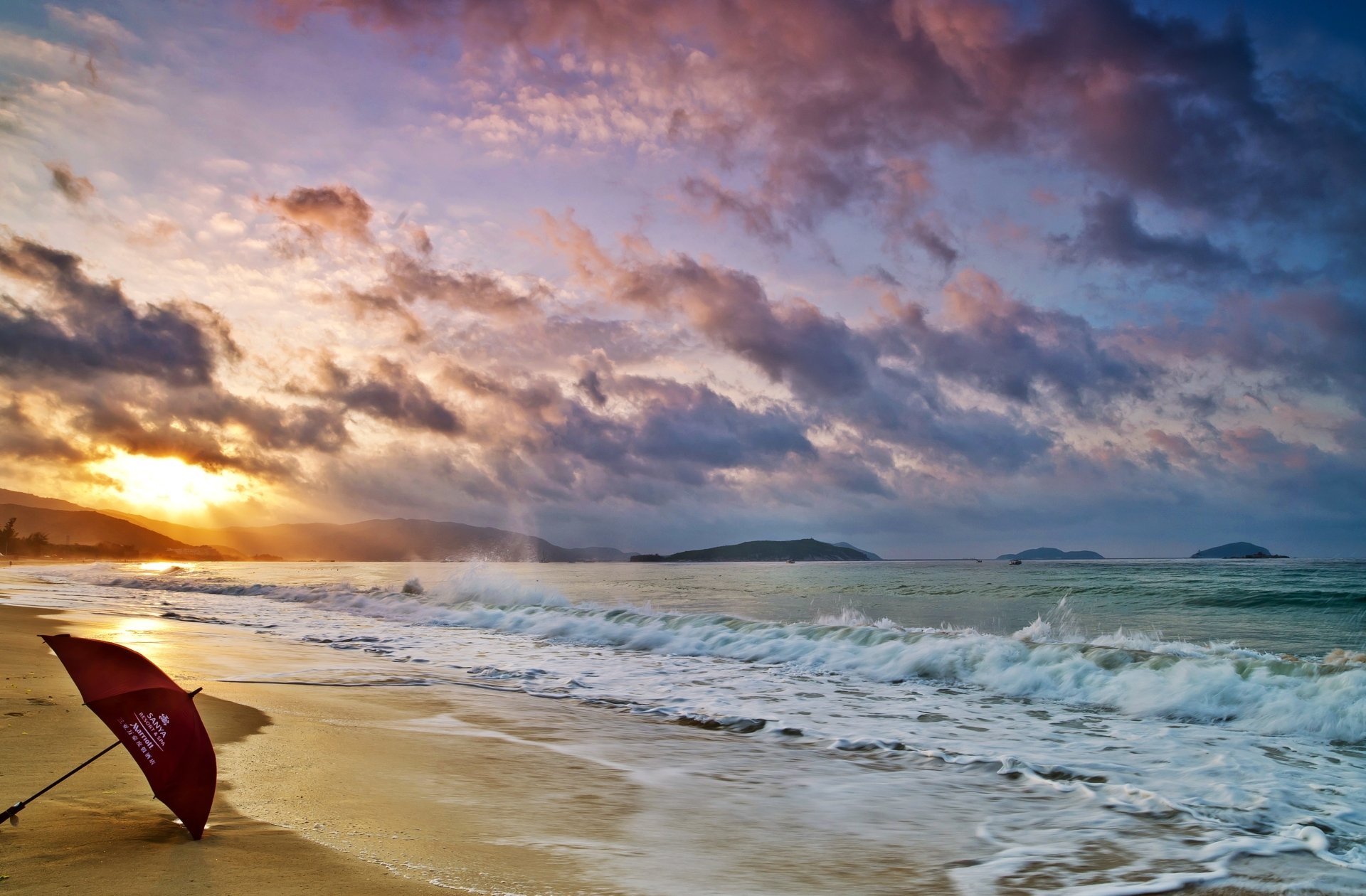 Sunrise over the ocean at a Hainan seashore in China, with waves gently crashing and a solitary umbrella on the sandy beach under a vibrant sky.