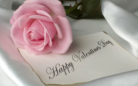 A pink rose rests on a silky fabric next to a card that reads Happy Valentine's Day, creating a beautiful Valentine's Day themed desktop wallpaper.