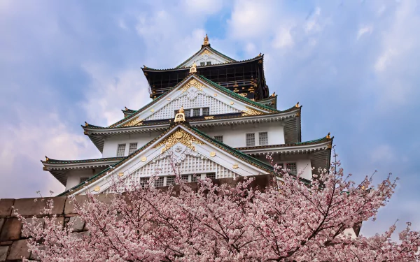 Osaka Castle framed by vibrant cherry blossom trees in full bloom during spring under a partly cloudy sky, captured in stunning 4K Ultra HD quality.