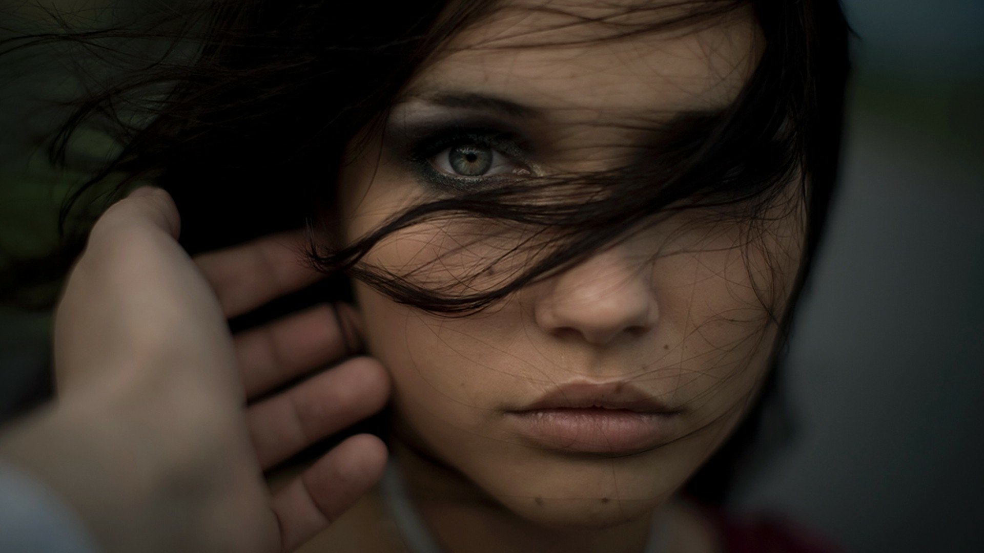 A close-up portrait of a brunette woman with captivating eyes, her face partially obscured by flowing hair, set against a blurred background. This HD image serves as a striking desktop wallpaper.