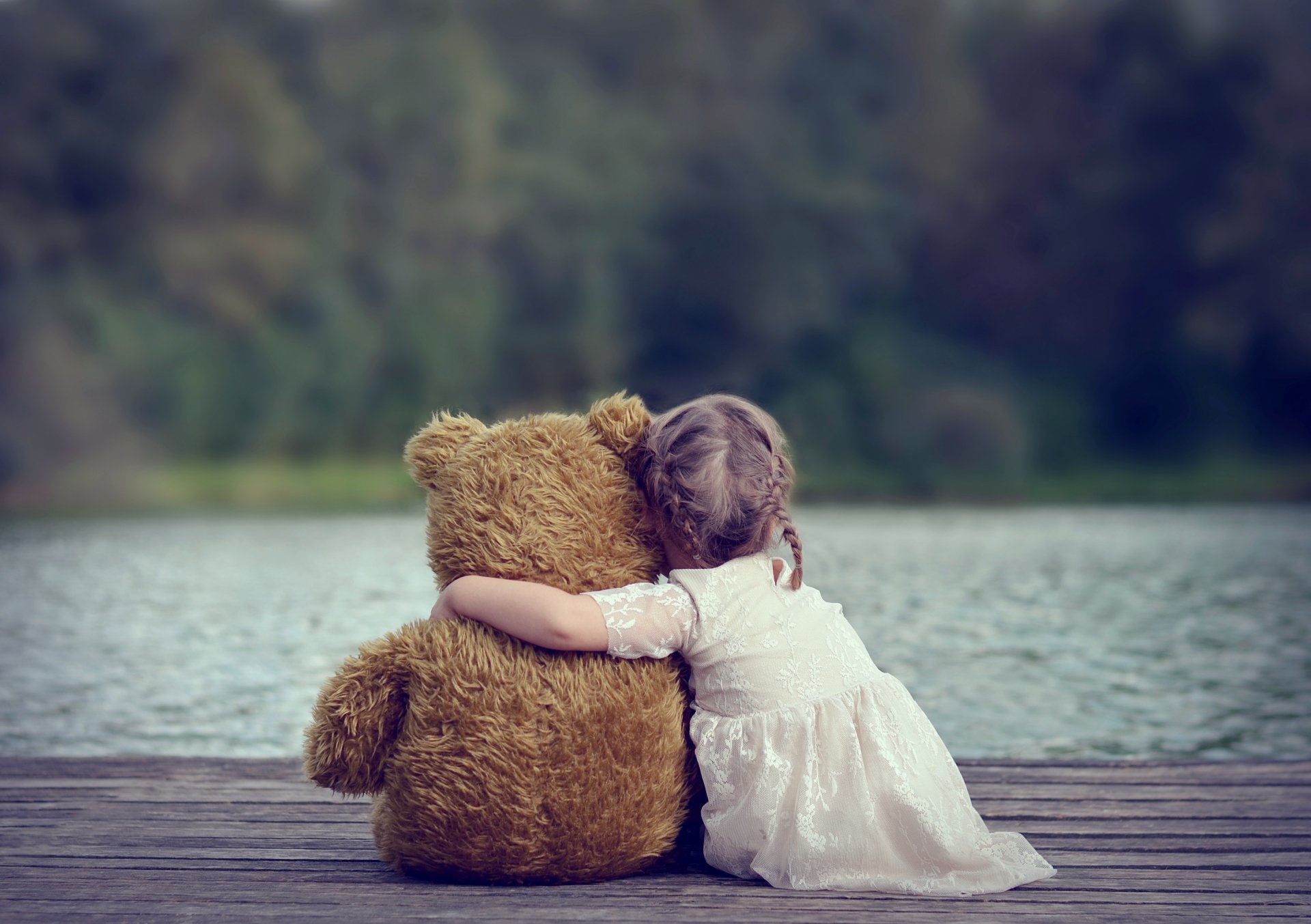 A child embraces a teddy bear while sitting on a dock by a serene lake, capturing a peaceful landscape in this 4K Ultra HD wallpaper.