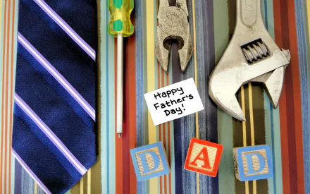 A colorful Father's Day desktop wallpaper featuring a tie, tools, and blocks spelling DAD, alongside a card that reads Happy Father's Day!