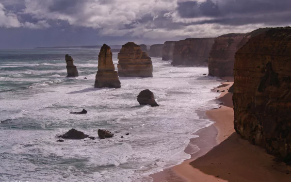 Dramatic ocean waves crash against limestone stacks along the Victoria coastline at The Twelve Apostles, Australia, beneath a cloudy sky in this 4K Ultra HD wallpaper.