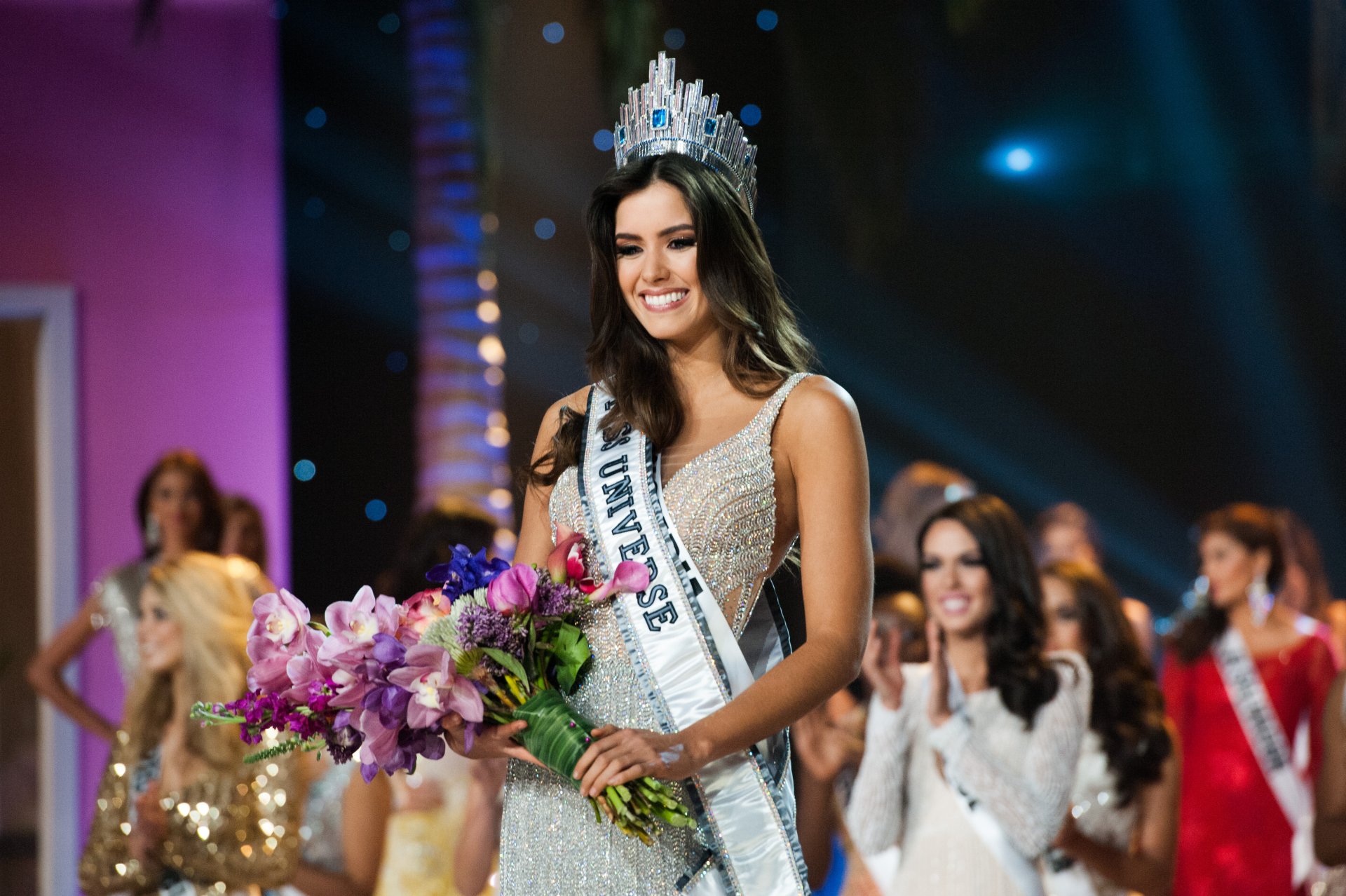 Paulina Vega, Miss Universe, smiles brightly while holding a vibrant bouquet of flowers, wearing her crown and sash. A captivating moment from the pageant, celebrating beauty and grace.