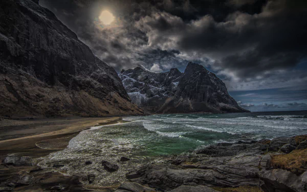 Dramatic coastline of Kvalvika, Lofoten Islands, Norway, features rugged mountains and turbulent seas under an atmospheric sky, creating a stunning natural landscape.