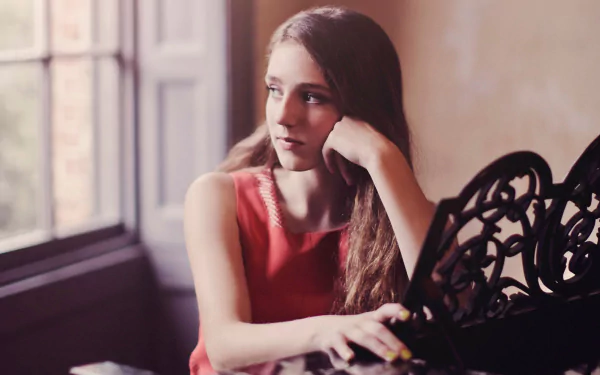 A HD wallpaper featuring Birdy, the English singer, contemplatively resting her chin on her hand while sitting near a window, capturing a moment of artistic reflection.