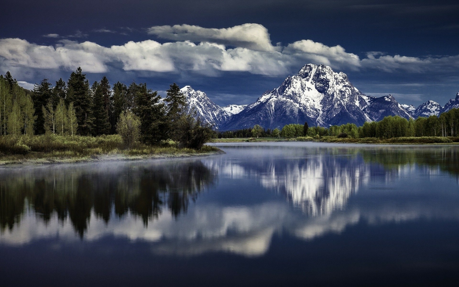 HD desktop wallpaper of Grand Teton National Park featuring snow-capped mountains, a forest, a river reflecting the cloudy sky, and serene natural beauty.