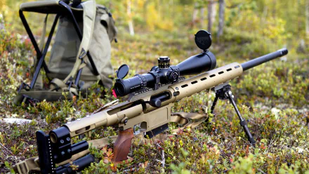 A detailed sniper rifle rests on the ground surrounded by greenery, with a backpack and chair in the background, providing a rugged outdoor scene for this HD desktop wallpaper.