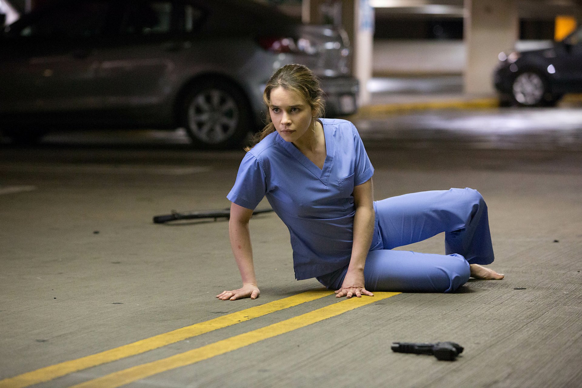 HD desktop wallpaper featuring Emilia Clarke in a scene from the movie Terminator Genisys, dressed in blue scrubs on a parking lot floor with a gun nearby.
