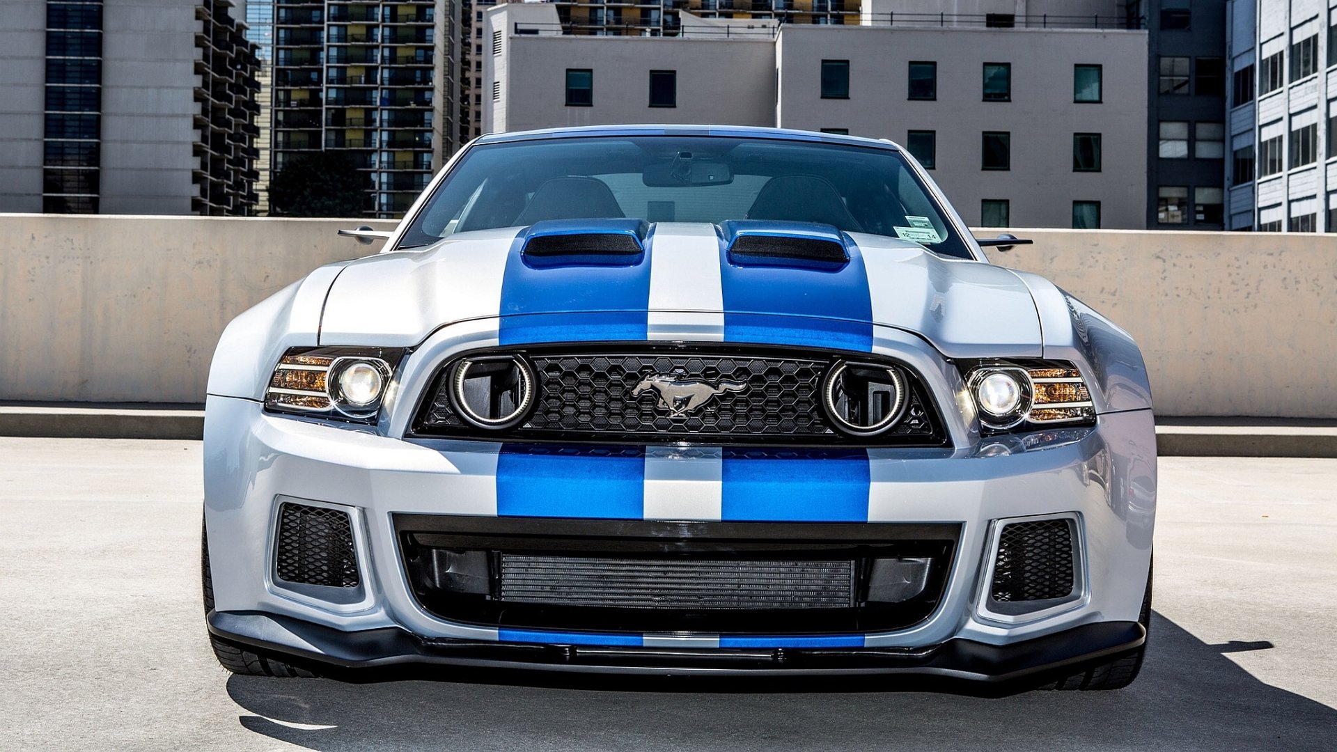 HD desktop wallpaper featuring a front view of a Ford Mustang, parked in an urban setting. The car has blue racing stripes on a silver body.