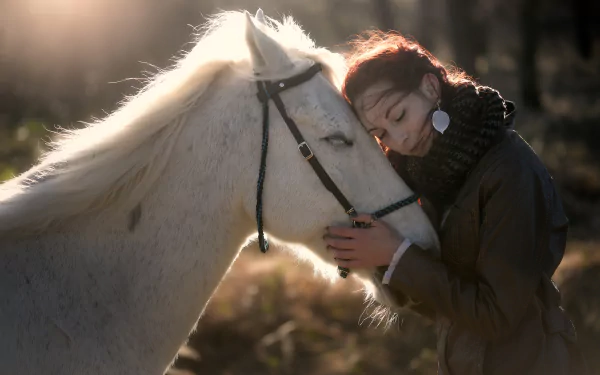 A serene HD wallpaper depicting a red-haired woman with earrings affectionately leaning her head against a white horse in a soft, diffused light, evoking a mood of love and tranquility.