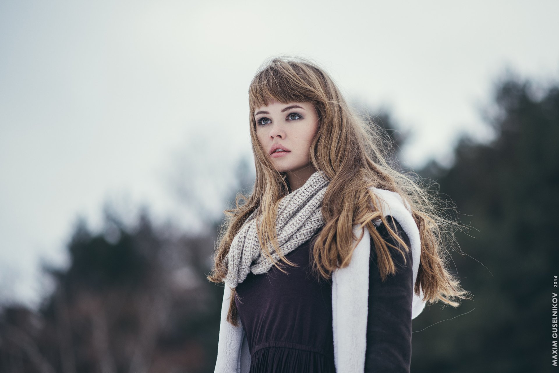 A model with brunette hair and green eyes wears a stylish scarf, standing in a serene outdoor setting, captured in HD as a striking desktop wallpaper.