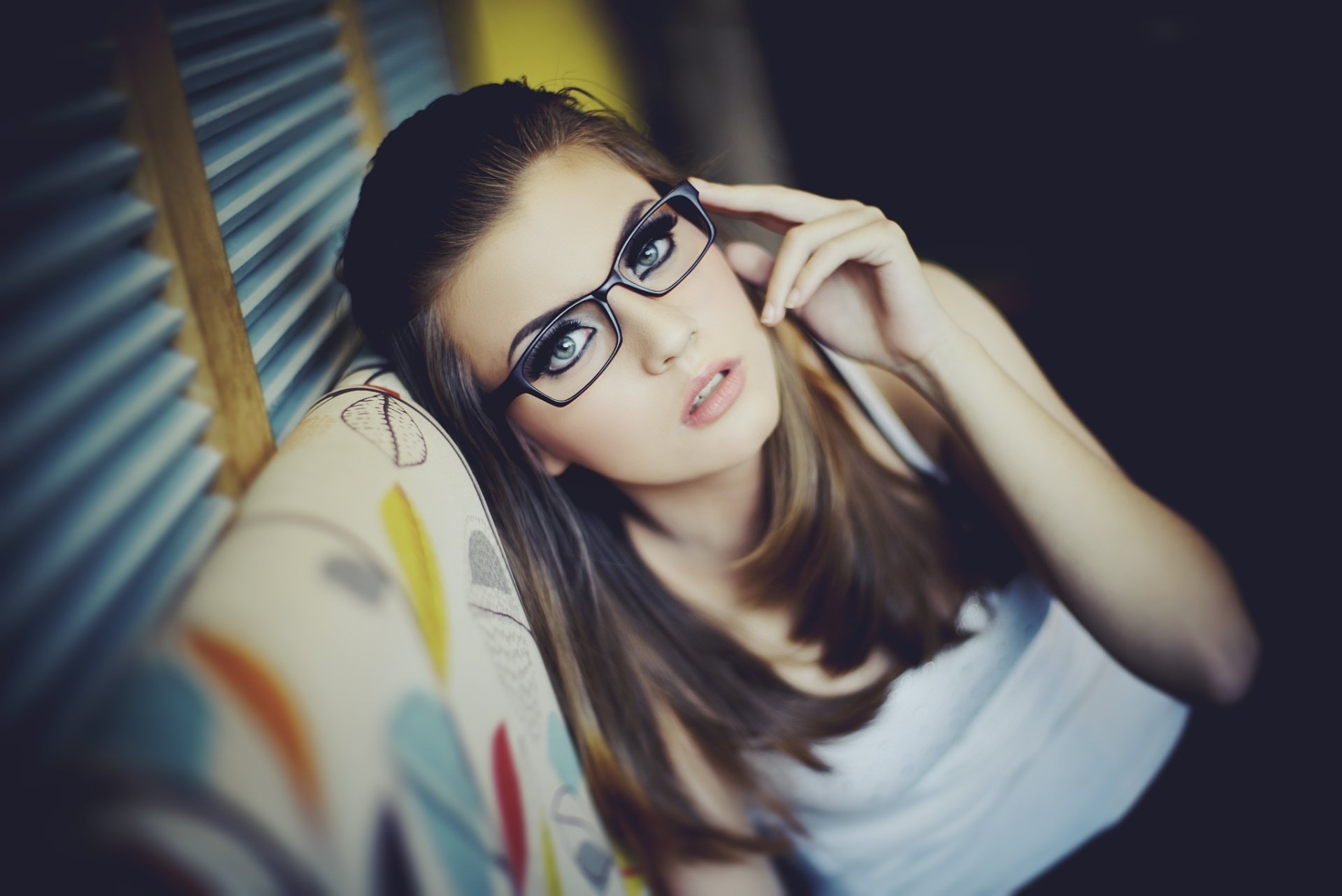 HD desktop wallpaper featuring a woman model with glasses, resting on a colorful couch, looking directly at the camera in soft, natural light.