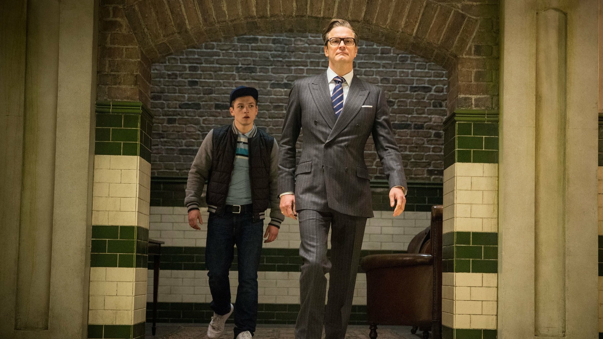 Taron Egerton and Colin Firth walk through a stylish corridor in a scene from Kingsman: The Secret Service. This image showcases their dynamic in the film.