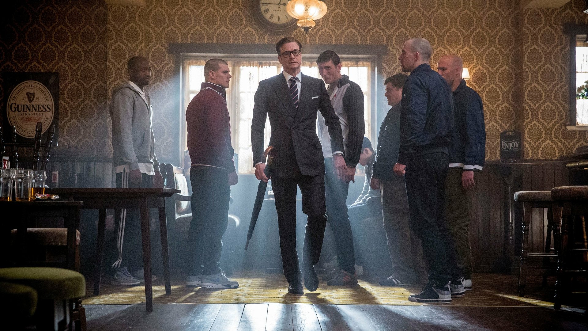 Colin Firth stands confidently at the center of a tense scene from Kingsman: The Secret Service, surrounded by a group of men in a vintage, dimly lit setting. A stylish 4K Ultra HD wallpaper.