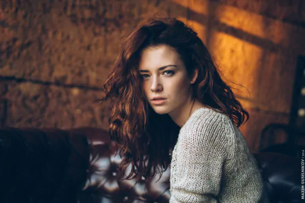 Red-haired woman with blue eyes, wearing a sweater, poses against a blurred background. The image features a warm, bokeh effect and serves as a high-definition desktop wallpaper.