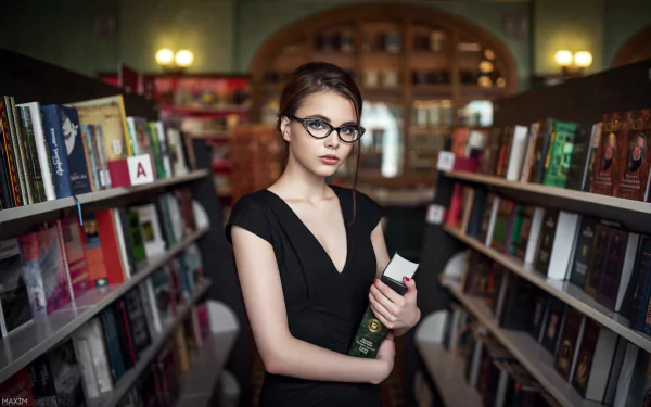 A brunette woman with brown eyes and glasses stands in a library, holding a book amidst shelves filled with colorful volumes, creating a bokeh effect in the background.