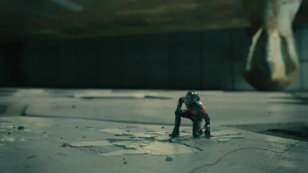 A dramatic HD wallpaper featuring Ant-Man in a crouching position on a cracked surface, framed by a shadowy, colossal object above, capturing a moment of intensity from the movie.
