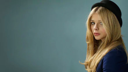 HD desktop wallpaper featuring celebrity Chloë Grace Moretz with long blonde hair and a black hat against a muted blue background.