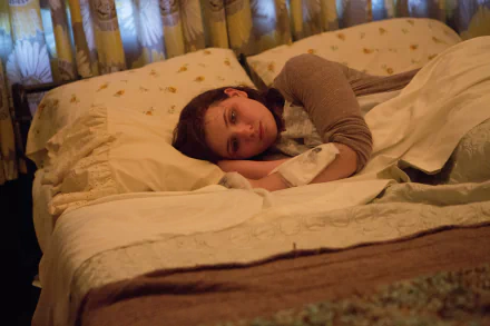 Still from the movie Maggie (2015): a young woman lies on a bed, gazing wistfully in warm bedroom light — HD PC desktop wallpaper/background.
