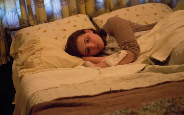 Still from the movie Maggie (2015): a young woman lies on a bed, gazing wistfully in warm bedroom light — HD PC desktop wallpaper/background.