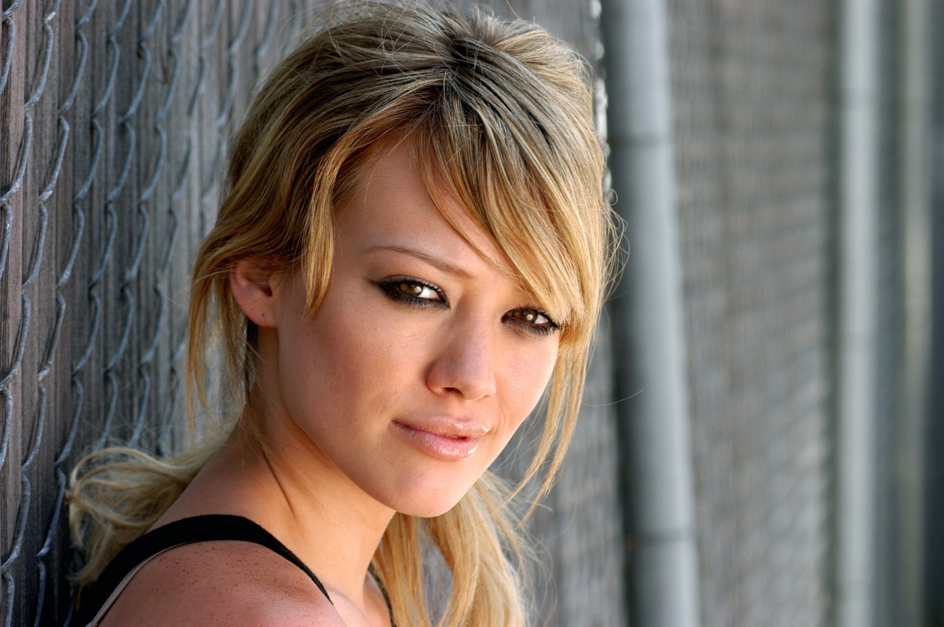 HD desktop wallpaper featuring a close-up of celebrity Hilary Duff with a soft expression, standing against a chain-link fence background.