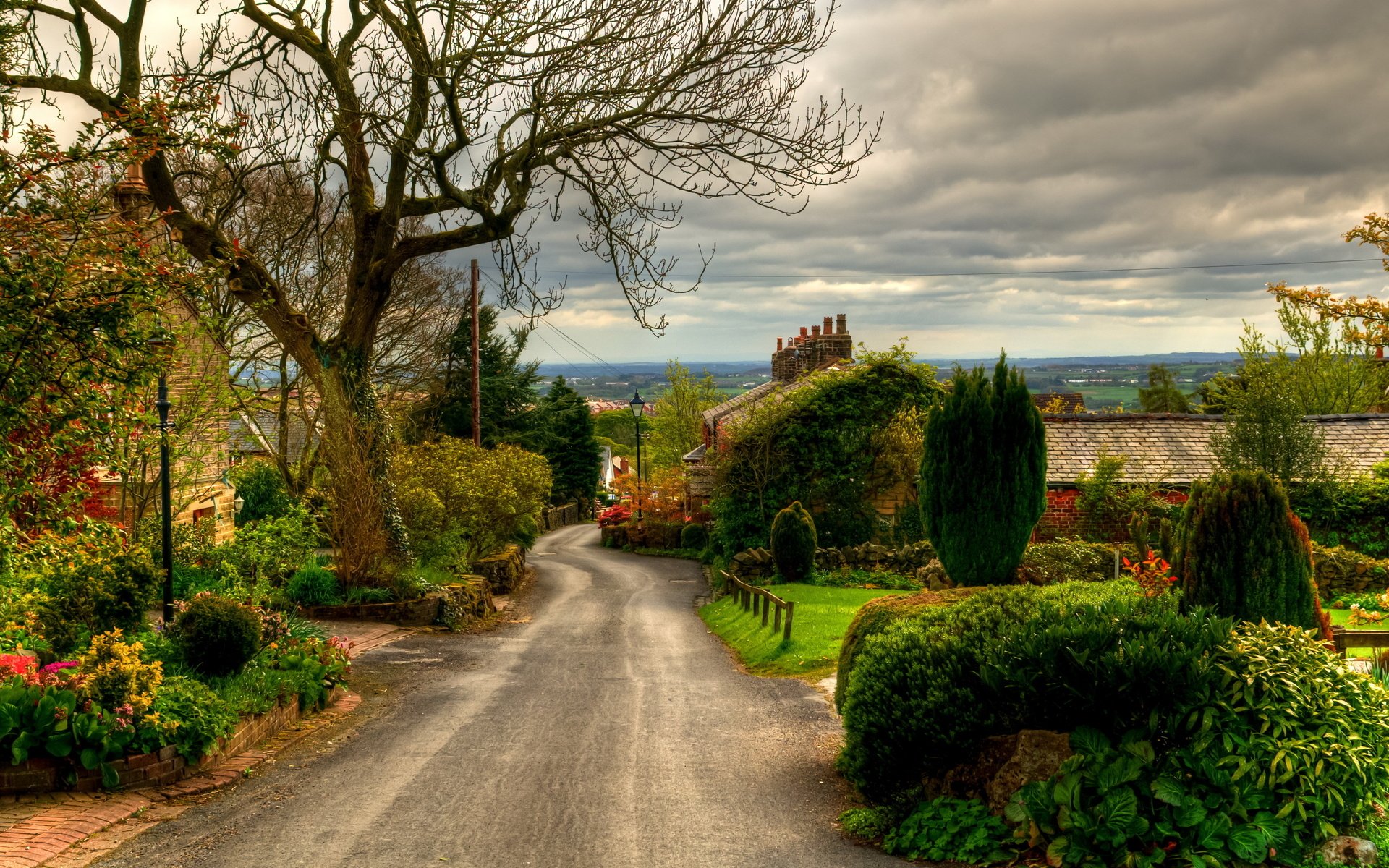 HD PC desktop wallpaper/background: a man-made country road winding past manicured gardens and cottages under a dramatic cloudy sky.