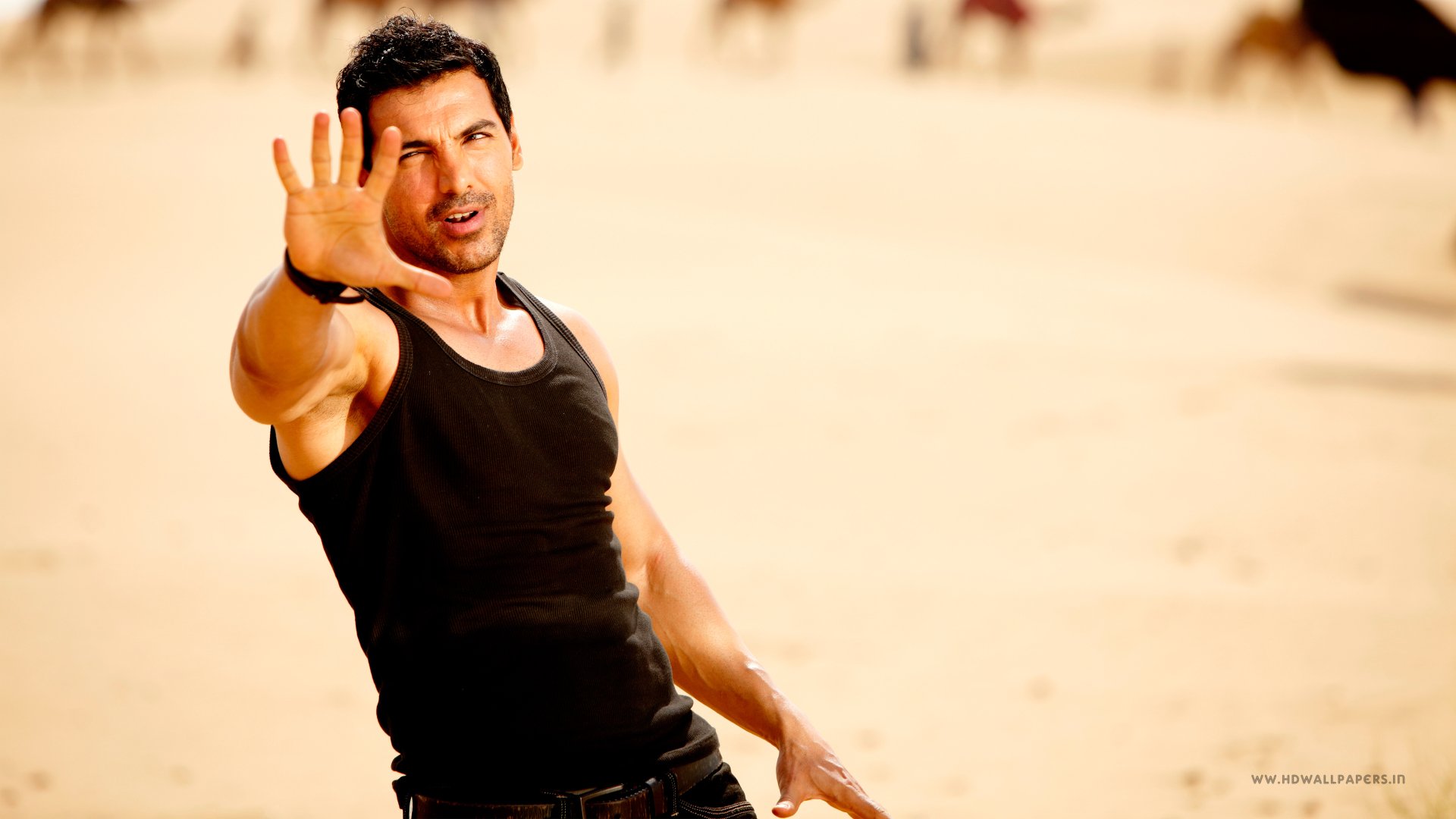 4K Ultra HD PC desktop wallpaper of a muscular man in a black tank top reaching toward the camera on a sunlit sandy beach.