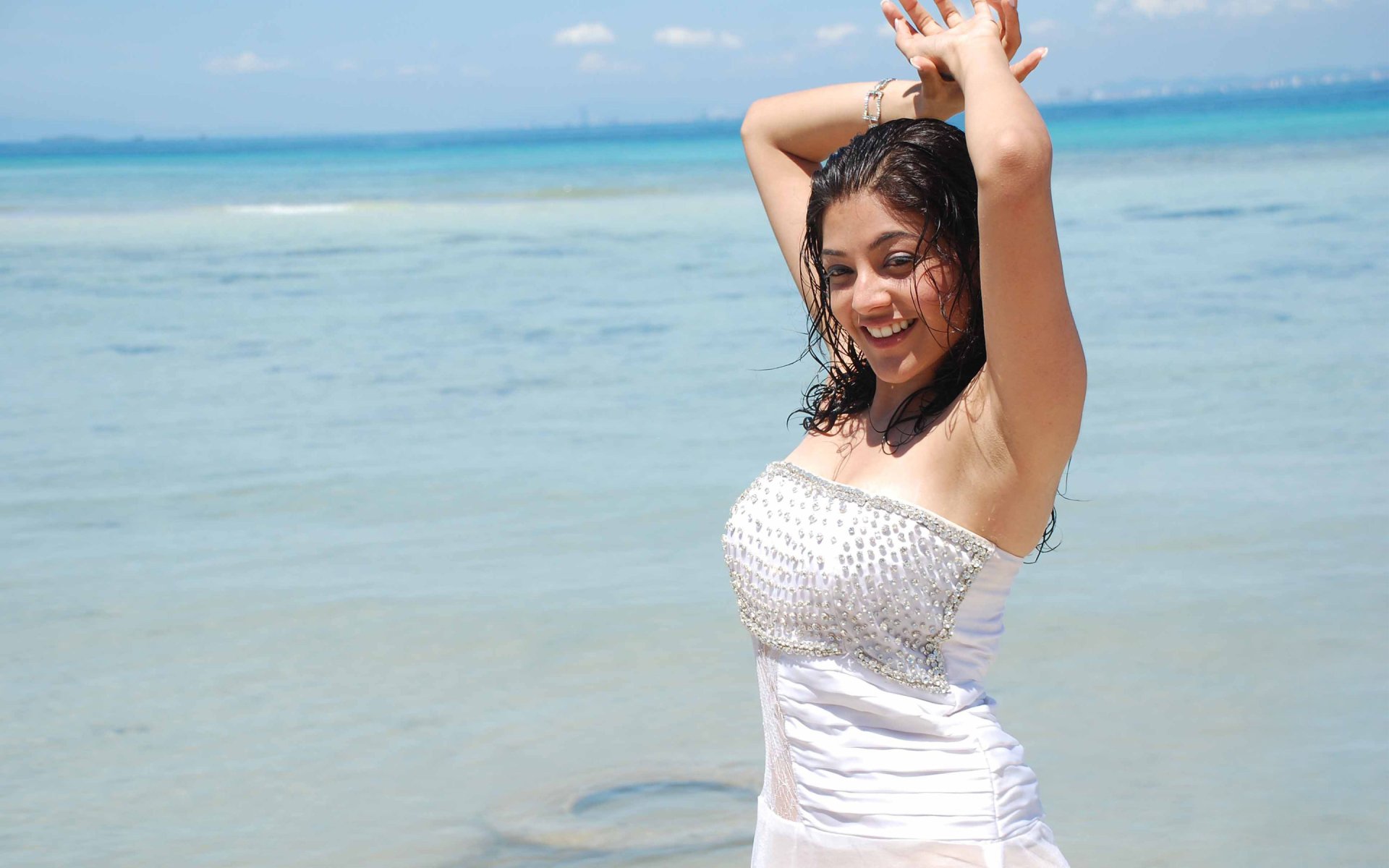 HD desktop wallpaper featuring celebrity Kajal Aggarwal, joyfully posing at the beach with a sparkling ocean backdrop. Her playful expression and elegant attire capture a vibrant summer vibe.