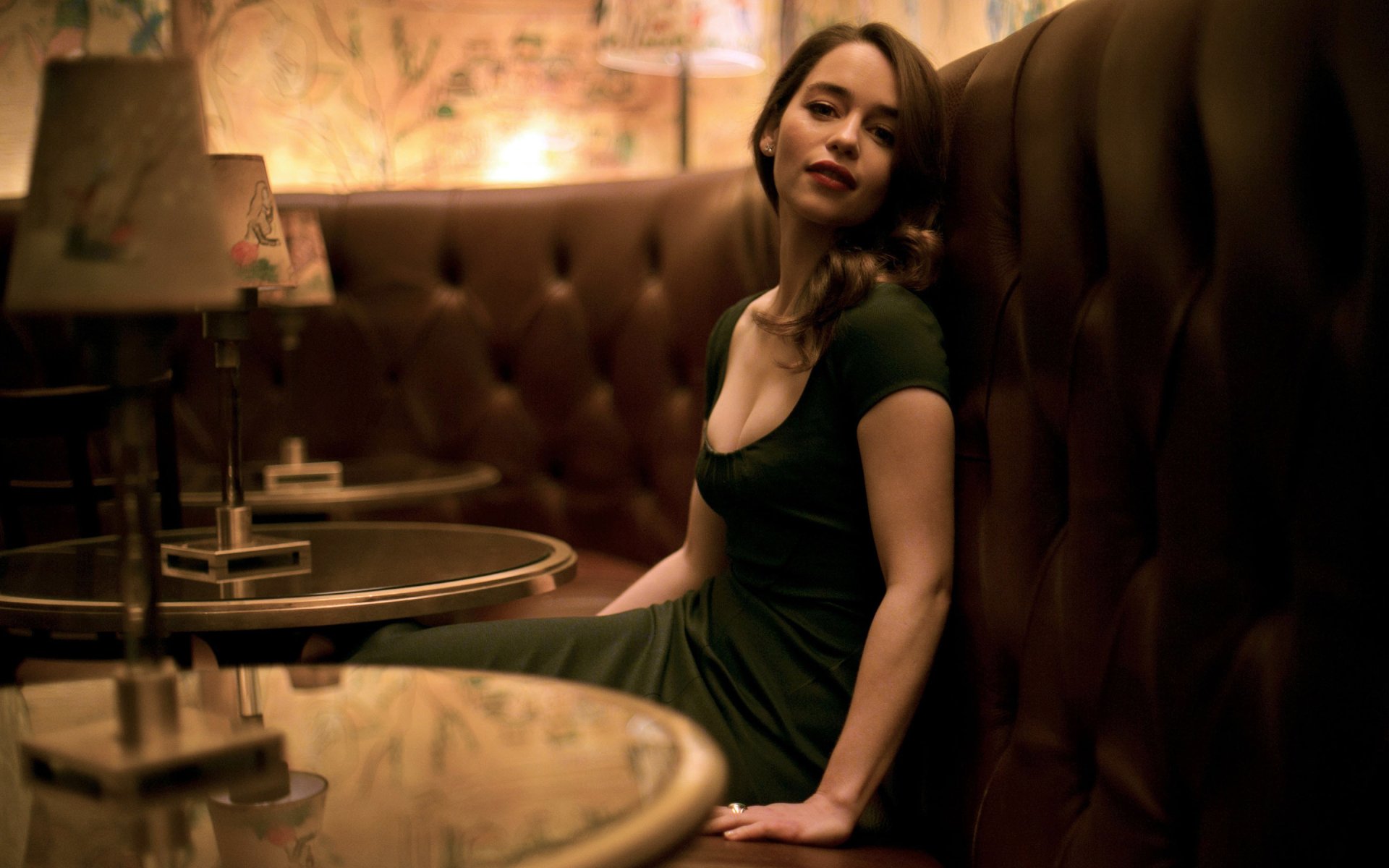 Emilia Clarke sits elegantly in a cozy, dimly lit lounge, wearing a green dress. The intimate setting features plush brown seating and stylish table lamps, creating a warm atmosphere.