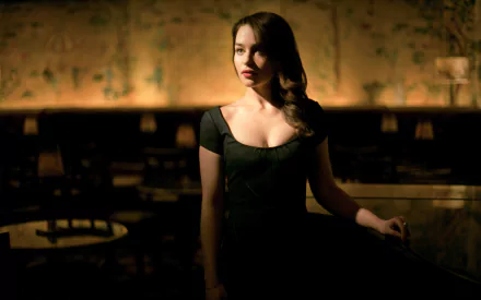 A striking HD wallpaper featuring Emilia Clarke, a brunette actress with long hair, posing elegantly in a dimly lit setting, exuding charm and sophistication.