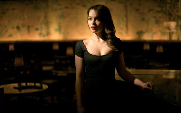 A striking HD wallpaper featuring Emilia Clarke, a brunette actress with long hair, posing elegantly in a dimly lit setting, exuding charm and sophistication.