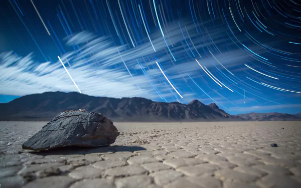HD nature desktop wallpaper showing star trails swirling over a barren desert with a large rock in the foreground and mountains in the distance.