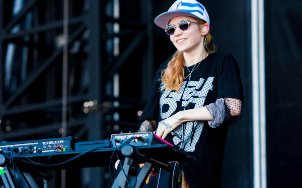 HD PC desktop wallpaper of music artist Grimes performing on stage with electronic equipment, wearing sunglasses and a cap.