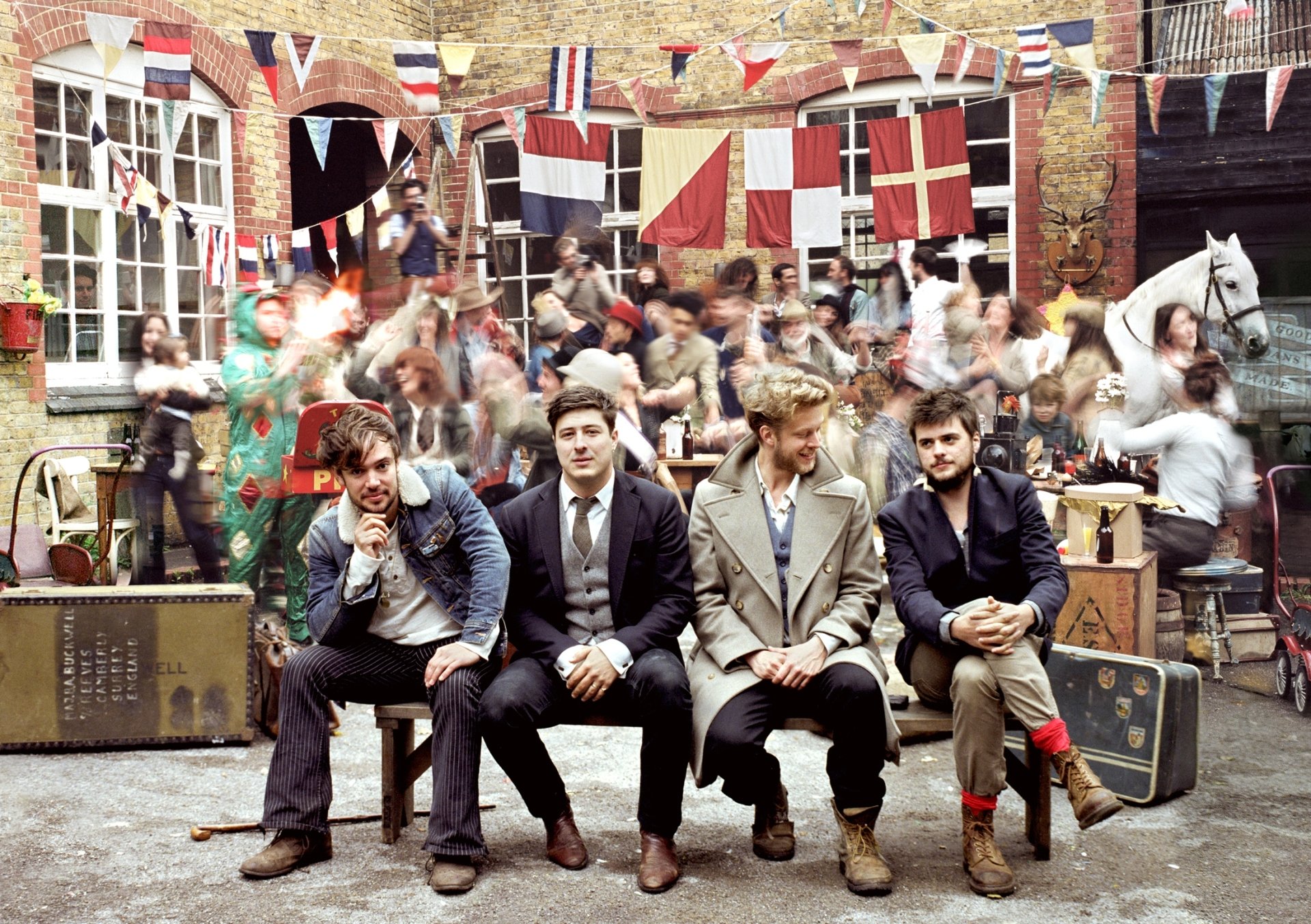 HD PC desktop wallpaper featuring Mumford & Sons seated in front of a lively courtyard scene with colorful flags, capturing the band's musical vibe and energy.