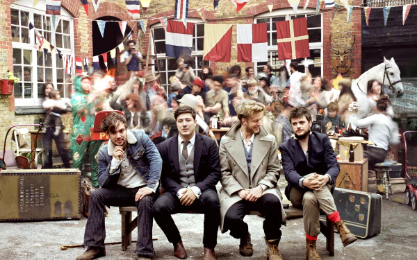 HD PC desktop wallpaper featuring Mumford & Sons seated in front of a lively courtyard scene with colorful flags, capturing the band's musical vibe and energy.