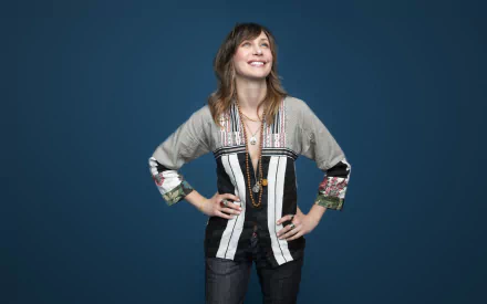 Vera Farmiga poses confidently against a solid blue background, wearing a stylish black and gray outfit. This image serves as a vibrant HD desktop wallpaper and background.