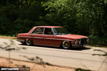 HD PC desktop wallpaper of a lowered red Mercedes‑Benz W114 parked on a sunlit roadside, framed by dense, leafy green trees.