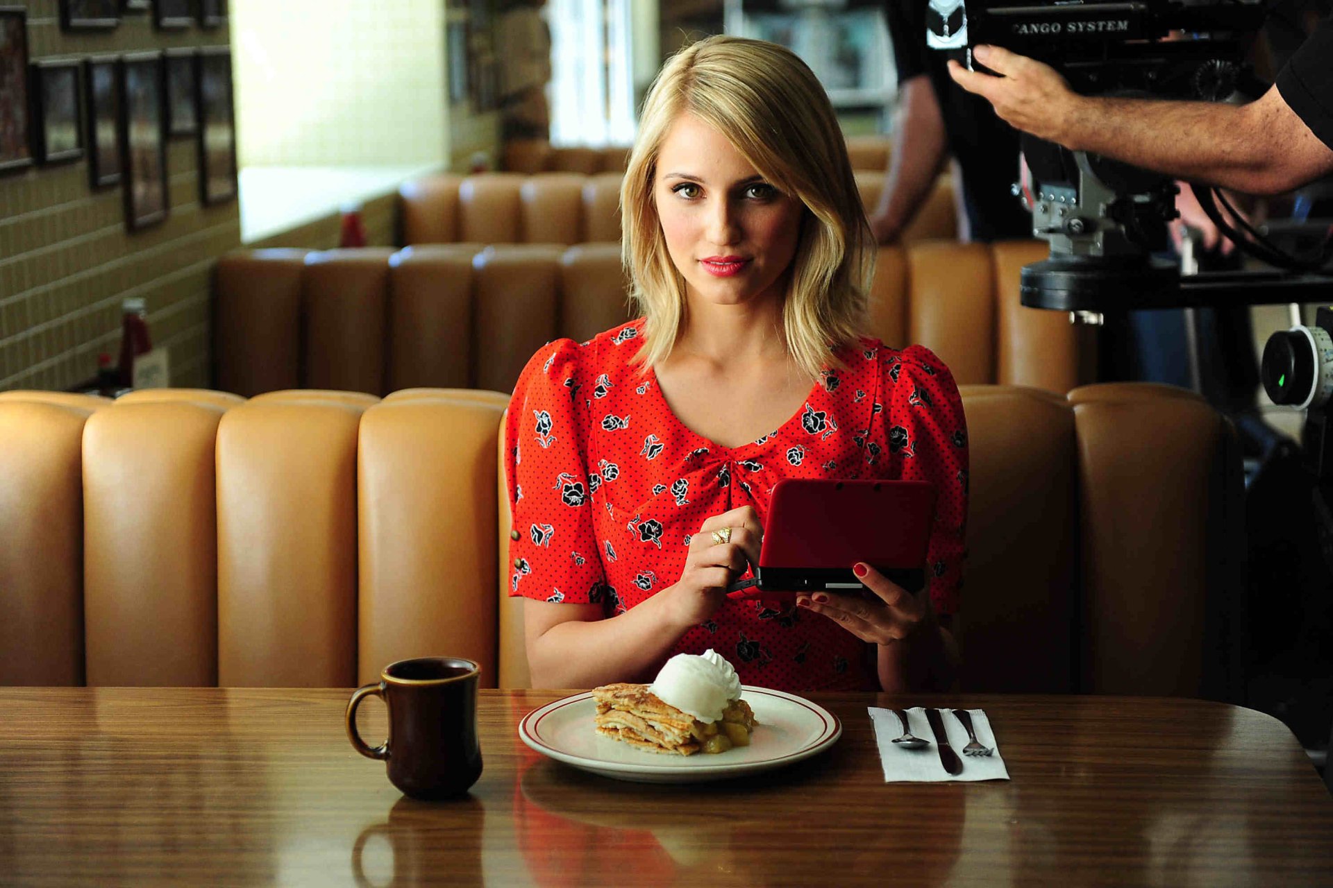 Dianna Agron sits at a diner table, holding a handheld gaming device, with a plate of food and a coffee cup in front of her. The vibrant scene serves as an engaging HD desktop wallpaper.