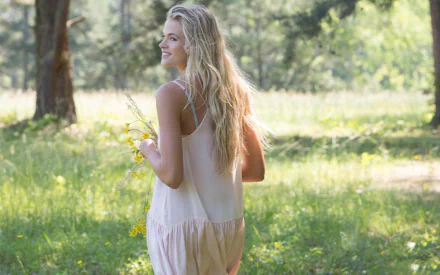 HD desktop wallpaper featuring Gabriella Wilde, a woman with long blonde hair, standing in a sunlit, grassy field holding yellow flowers. She is wearing a light, sleeveless dress and looking over her shoulder.