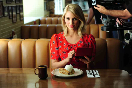 Dianna Agron sits at a diner table, holding a handheld gaming device, with a plate of food and a coffee cup in front of her. The vibrant scene serves as an engaging HD desktop wallpaper.