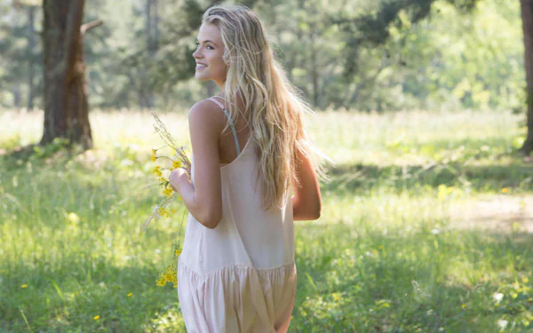 HD desktop wallpaper featuring Gabriella Wilde, a woman with long blonde hair, standing in a sunlit, grassy field holding yellow flowers. She is wearing a light, sleeveless dress and looking over her shoulder.